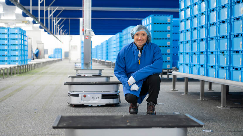 Monika Hoektra, Programmadirecteur nxtgen, gehurkt naast een AMR-robot in een groentefabriek met stapels blauwe kratten
